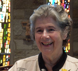 Photograph of a woman with gray hair, standing in front of stained glass windows